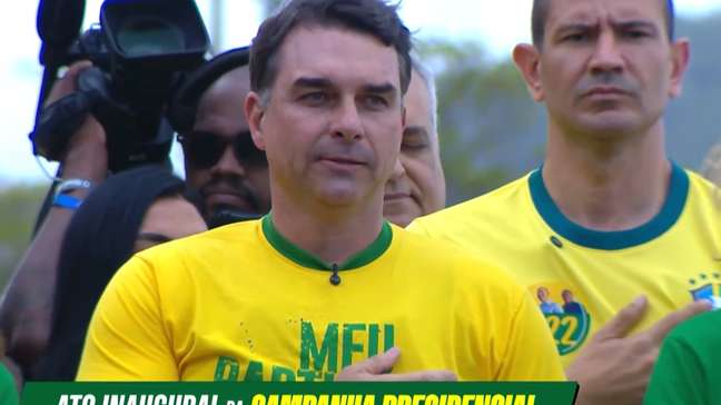 Flávio Bolsonaro lança campanha com colete à prova de balas e canta hino ao lado da mãe e da esposa